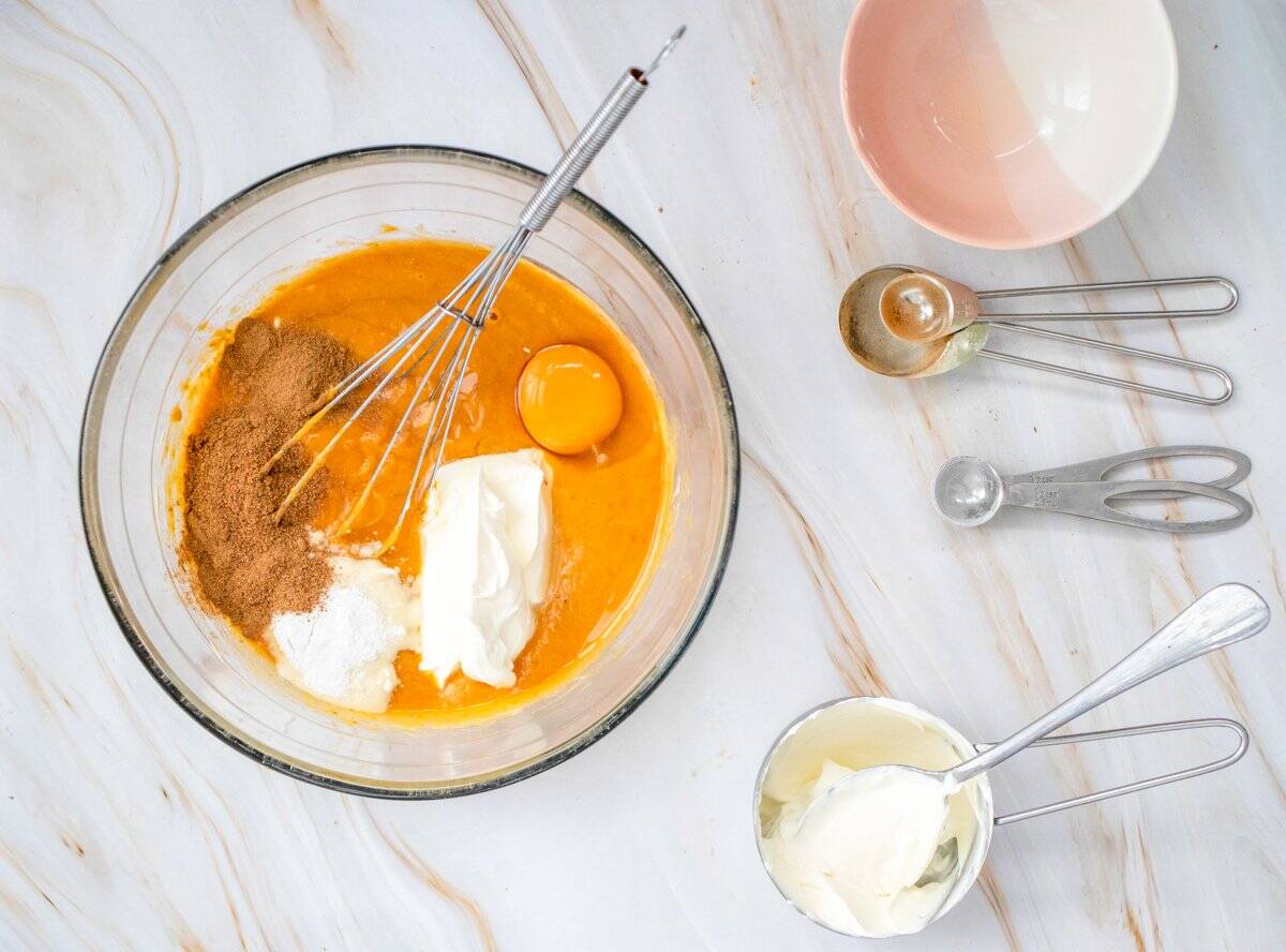 A glass bowl contains a mixture of pumpkin puree, eggs, and brown sugar, surrounded by measuring spoons filled with spices and a cup of sour cream, suggesting preparation for an easy pumpkin crisp recipe.
