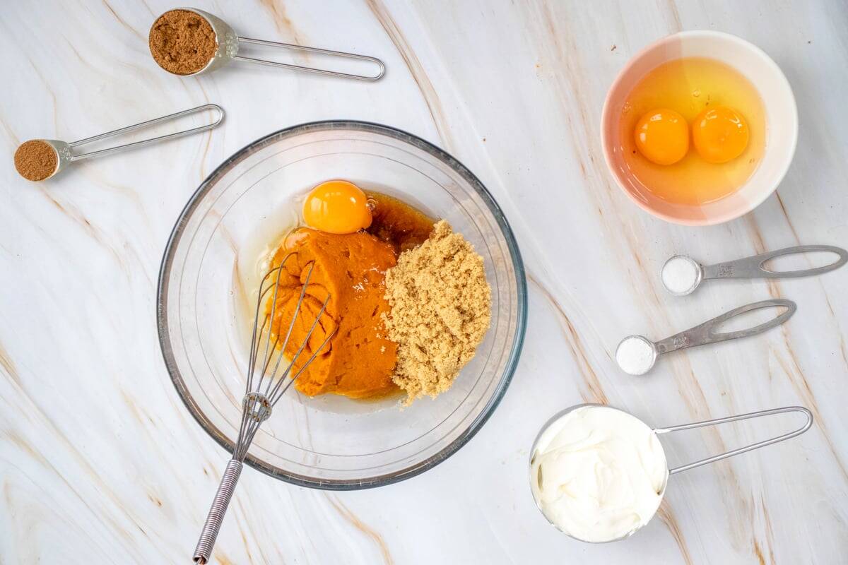A glass bowl contains a mixture for pumpkin crisp, featuring pumpkin puree, an egg, sugar, spices, and cream cheese, alongside measuring spoons and a small bowl with additional ingredients.
