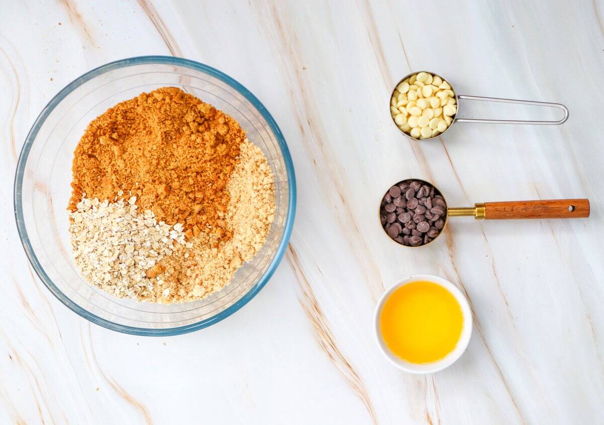 A glass bowl filled with a mixture of brown sugar, oats, and crumbs is surrounded by measuring cups containing white chocolate chips, chocolate chips, and melted butter, highlighting ingredients for an easy pumpkin crisp recipe.
