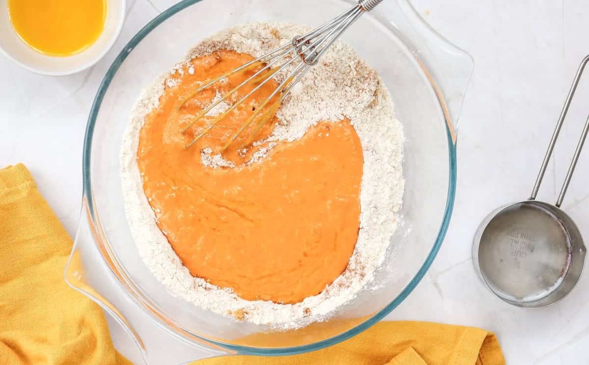 A glass mixing bowl contains a mixture of pumpkin puree and flour, with a whisk resting inside, accompanied by a bowl of eggs, a measuring cup, and a yellow cloth, suggesting preparation for an easy pumpkin pancake recipe.