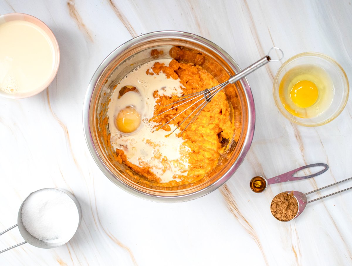 A stainless steel bowl containing pumpkin puree, eggs, sugar, and a whisk is surrounded by measuring cups with ingredients for an easy pumpkin dump cake recipe on a marble surface.
