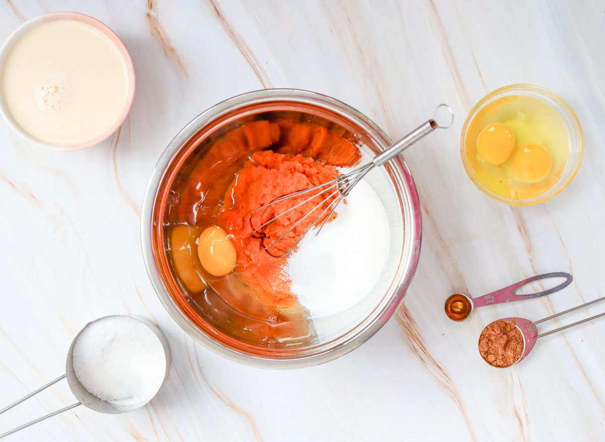 A metal mixing bowl contains pumpkin puree alongside measuring cups of sugar, pumpkin spice, and eggs, arranged on a marble countertop, suggesting the preparation of an easy pumpkin dessert recipe.
