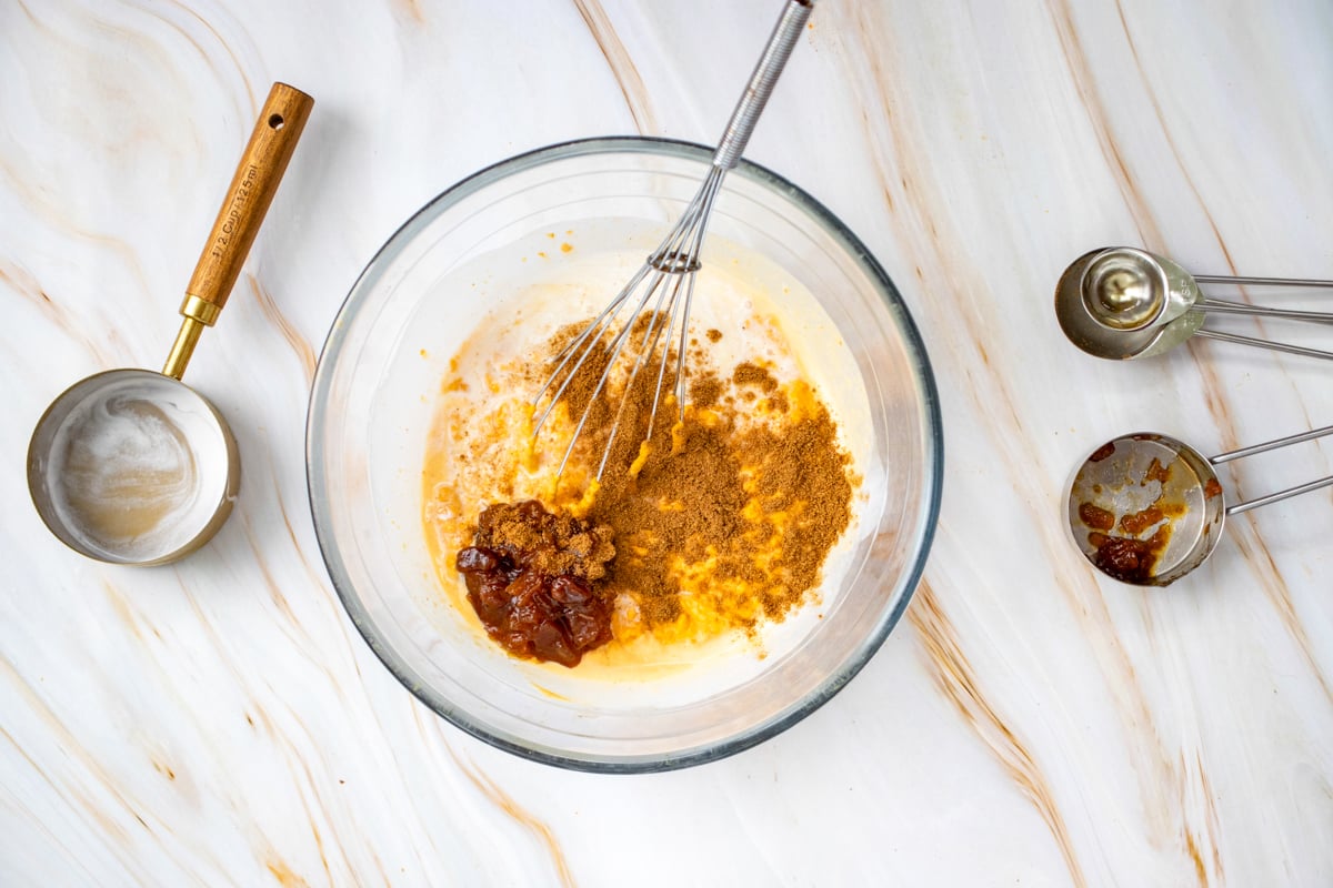 A glass bowl contains a mixture of pumpkin puree and sugar, surrounded by measuring cups of brown sugar, vanilla extract, and a whisk on a marble countertop, illustrating ingredients for a gluten free pumpkin dump cake.
