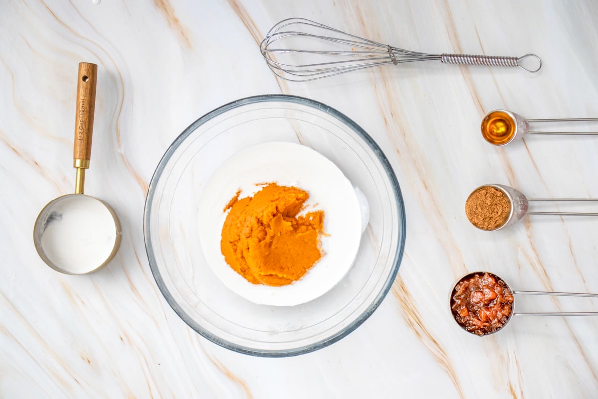 A glass mixing bowl contains a whisked mixture of pumpkin puree, brown sugar, and other ingredients on a marble surface, with measuring spoons and a cup nearby, suggesting preparation for a gluten free pumpkin dump cake or dessert.
