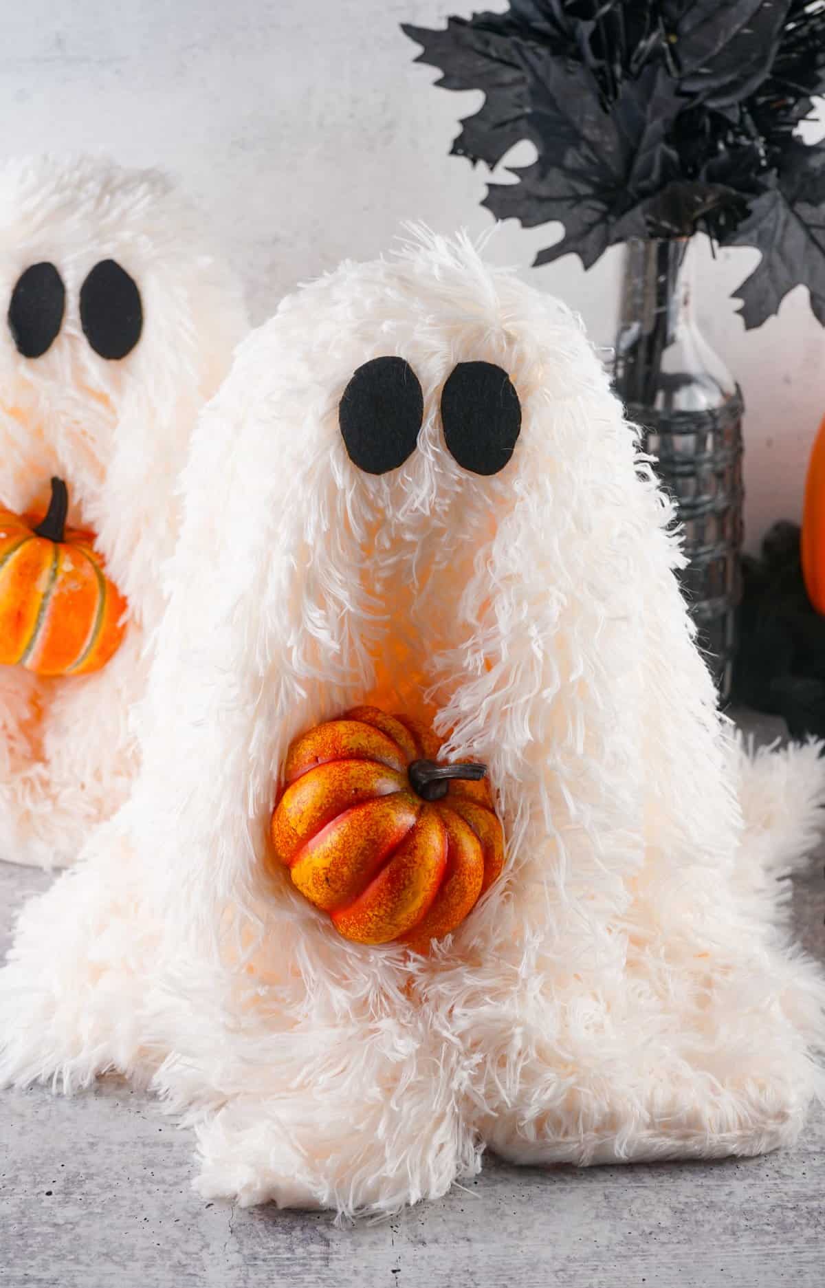Two fluffy white DIY ghosts hold orange pumpkins, set against a backdrop of fall decor, embodying a cozy Halloween DIY theme.
