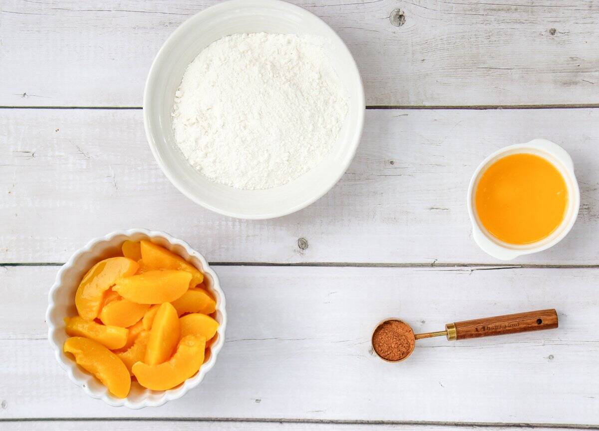 Ingredients for a fresh peach dump cake recipe are displayed, including a bowl of sliced peaches, a plate of flour, a bowl of melted butter, and a measuring spoon of cinnamon on a wooden surface.