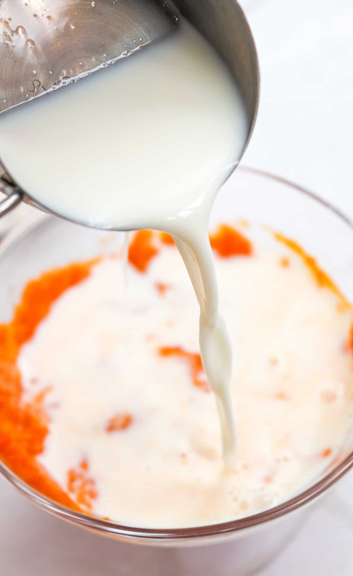 A stream of milk is being poured into a bowl containing orange puree, likely as part of a quick pumpkin pancake recipe for fluffy pumpkin pancakes or pumpkin spice pancakes.