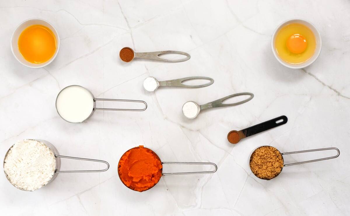 An overhead view of ingredients for a pumpkin pancake recipe, including eggs, milk, flour, pumpkin puree, brown sugar, and spices, arranged on a marble surface.