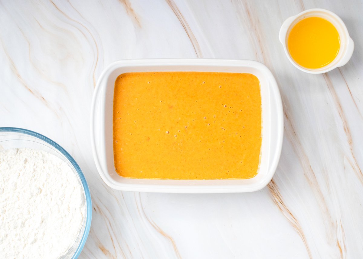 A white baking dish filled with a smooth, orange pumpkin mixture sits next to a bowl of flour and a small bowl of melted butter on a marble countertop, illustrating the preparation for an easy pumpkin dump cake recipe.
