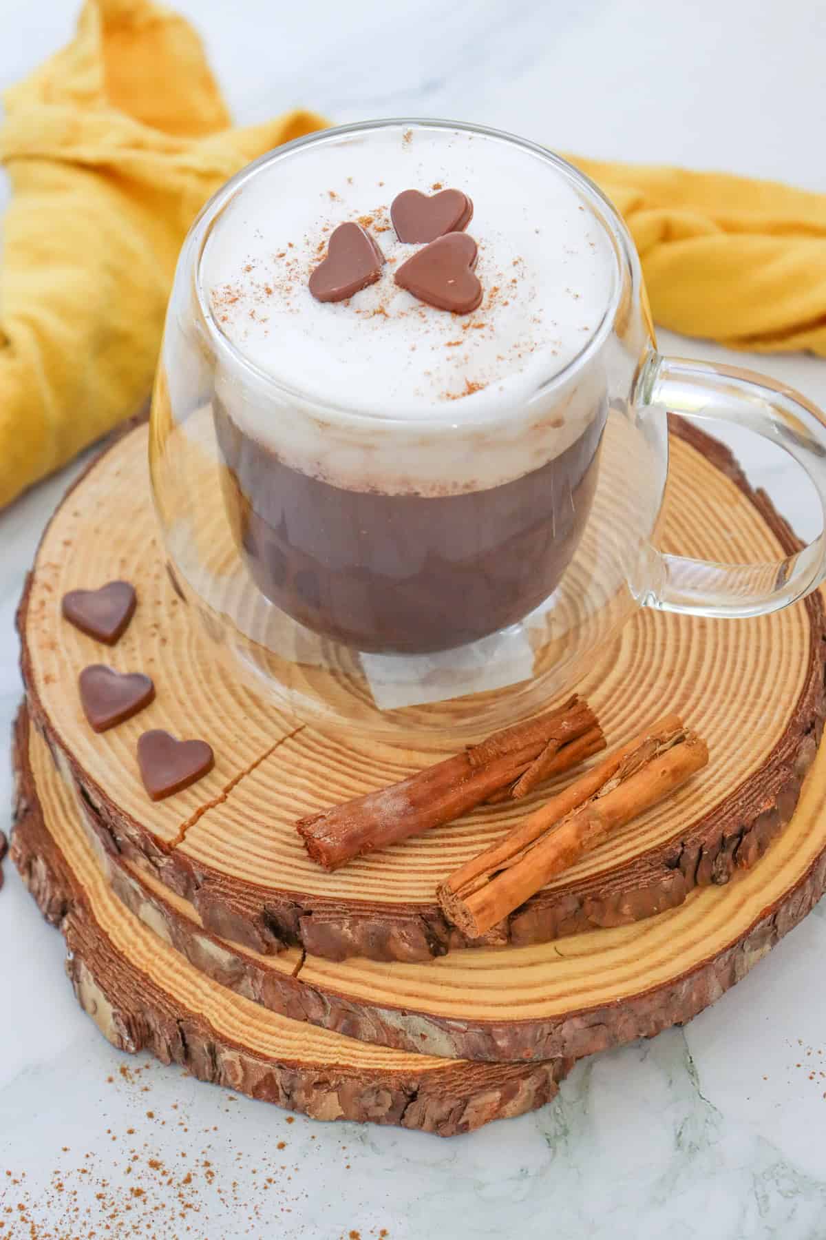 A clear glass mug filled with hot chocolate topped with whipped cream, chocolate heart candies, and a sprinkle of cinnamon sits on a wooden slice, accompanied by cinnamon sticks and a yellow cloth in the background, illustrating ideas for fall hot chocolate recipes such as pumpkin spice hot chocolate.
