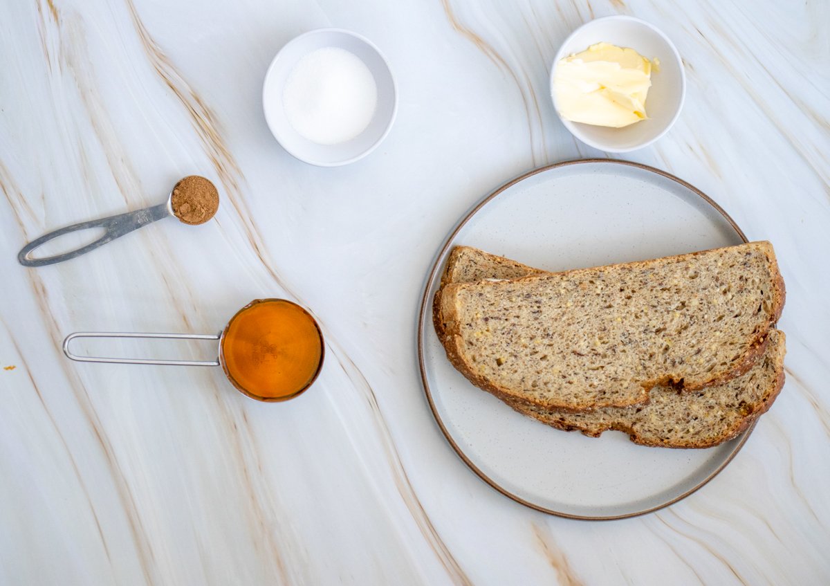A plate holding two slices of sourdough toast is accompanied by a small bowl of butter, a bowl of sugar, a measuring spoon of cinnamon, and a jar of maple syrup on a marble surface, illustrating potential toppings for sourdough toast recipes such as sweet and breakfast options.
