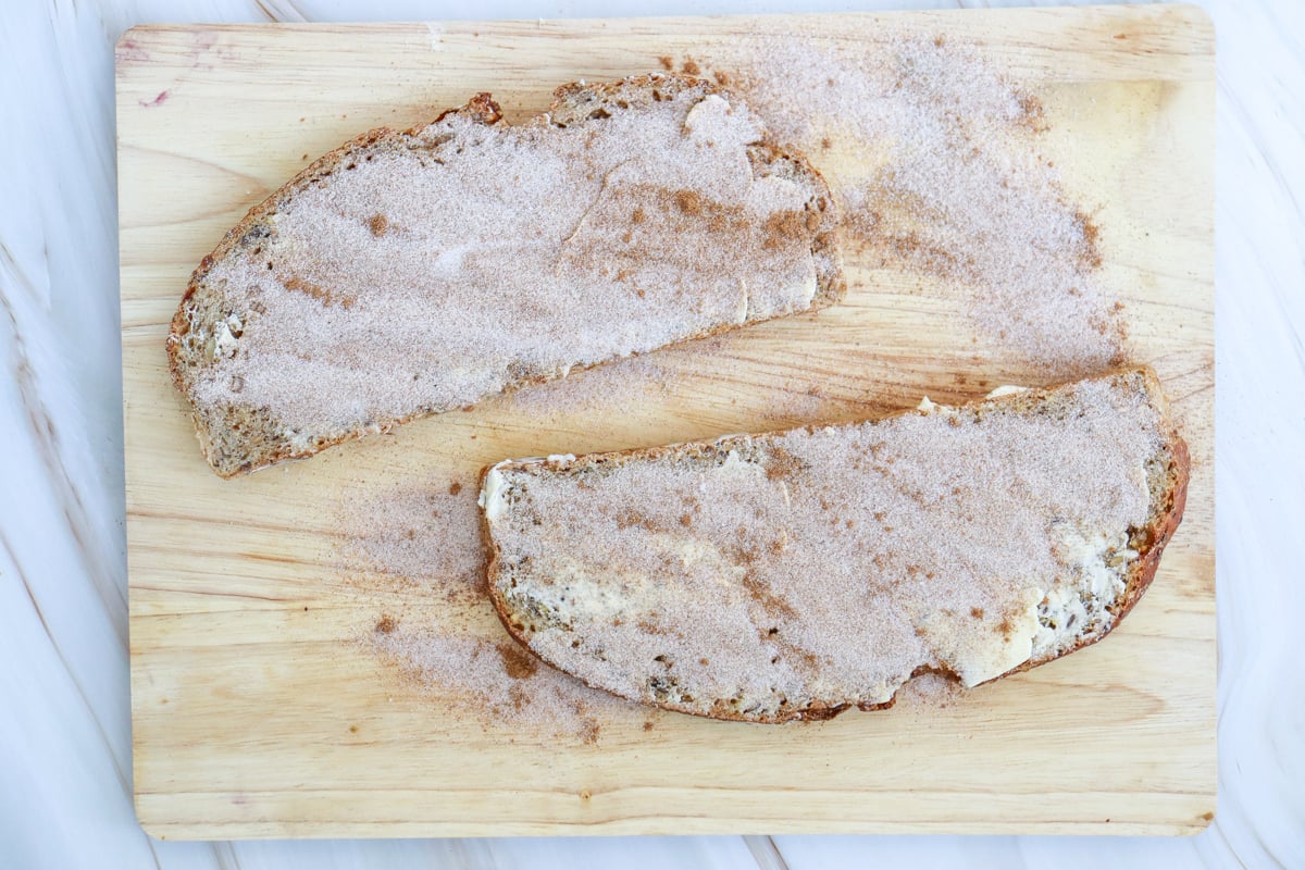 Two slices of sourdough toast, topped with a mixture of cinnamon and sugar, are placed on a wooden cutting board.
