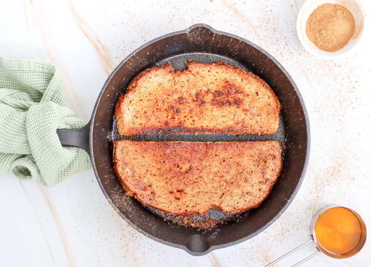 A cast iron skillet holds two slices of toasted sourdough bread, accompanied by a small dish of brown sugar and a measure of syrup, suggesting ideas for sweet sourdough toast toppings.
