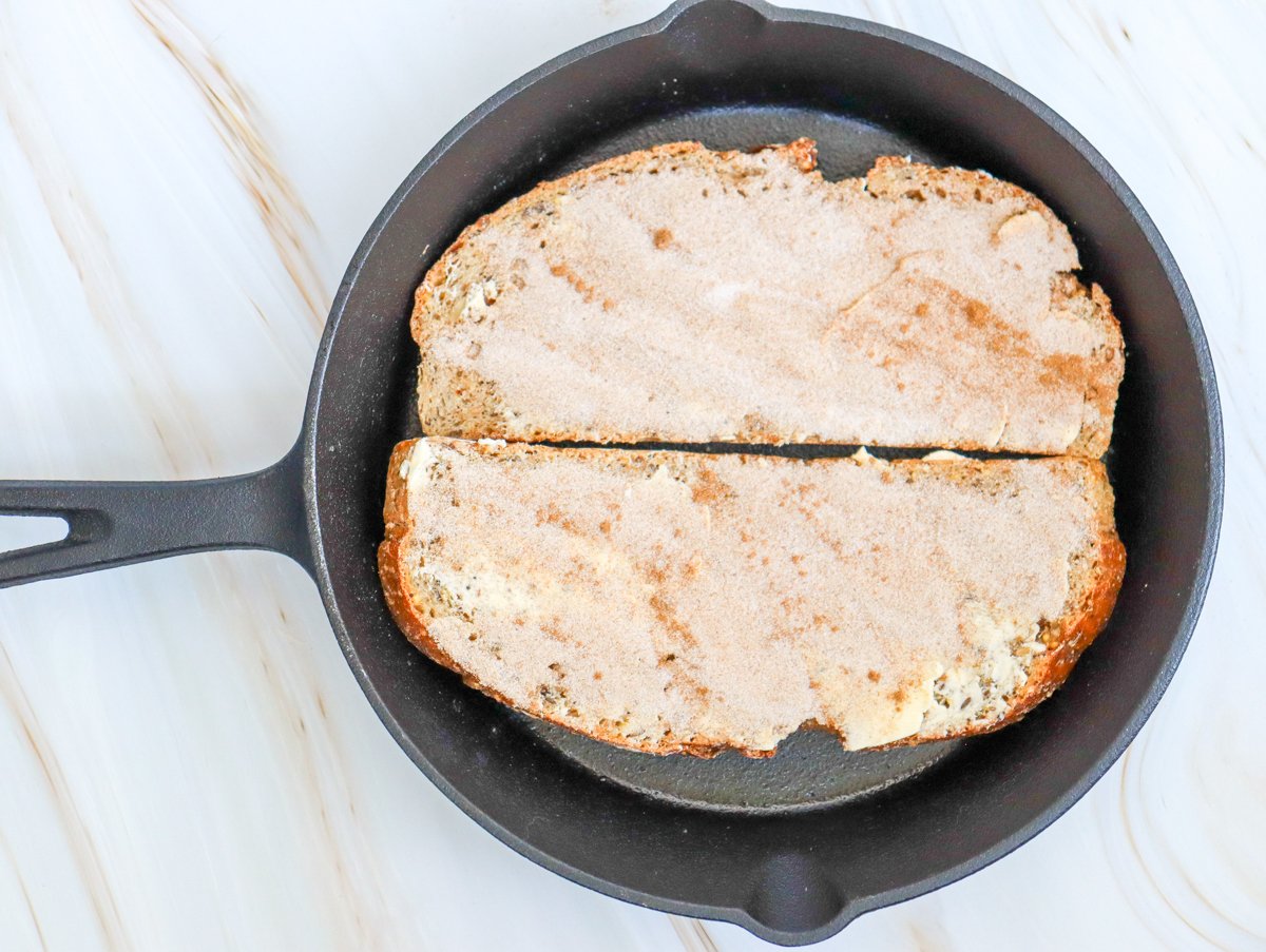 A cast iron skillet holds two slices of toasted sourdough bread, prepared with a layer of butter and a sprinkle of cinnamon sugar.
