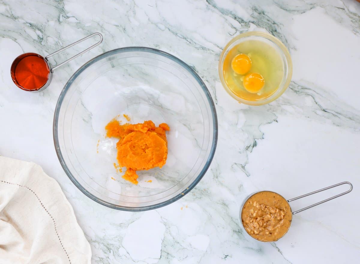 A glass bowl contains a mixture of mashed sweet potatoes, while a separate bowl holds two eggs and a measuring cup filled with peanut butter