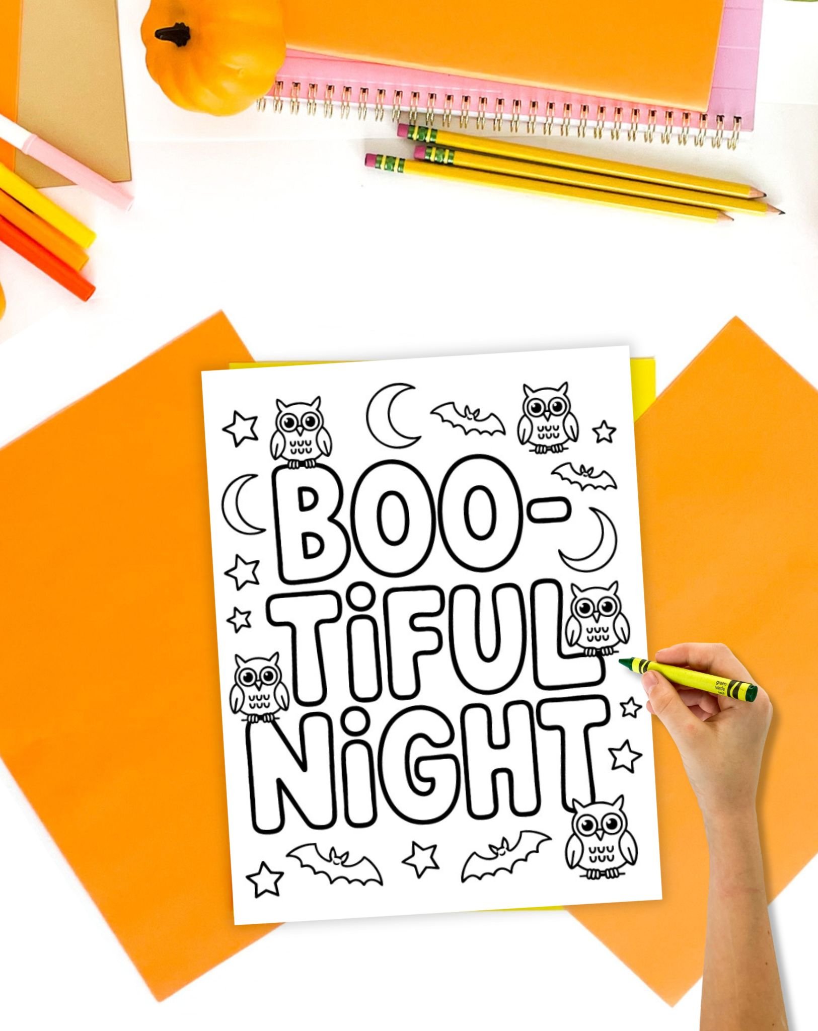 A hand holding a green crayon colors in a "Boo-tiful Night" Halloween-themed printable coloring page featuring owls, bats, and stars, surrounded by colorful stationery and decorations for Halloween activities.
