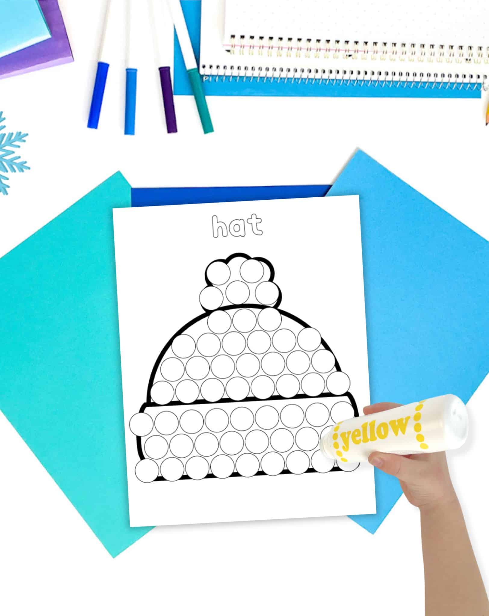 A hand is holding a yellow dot marker over a printable activity sheet featuring a winter hat outline, surrounded by colored papers and markers, ideal for preschool winter holiday activities with do a dot printables.
