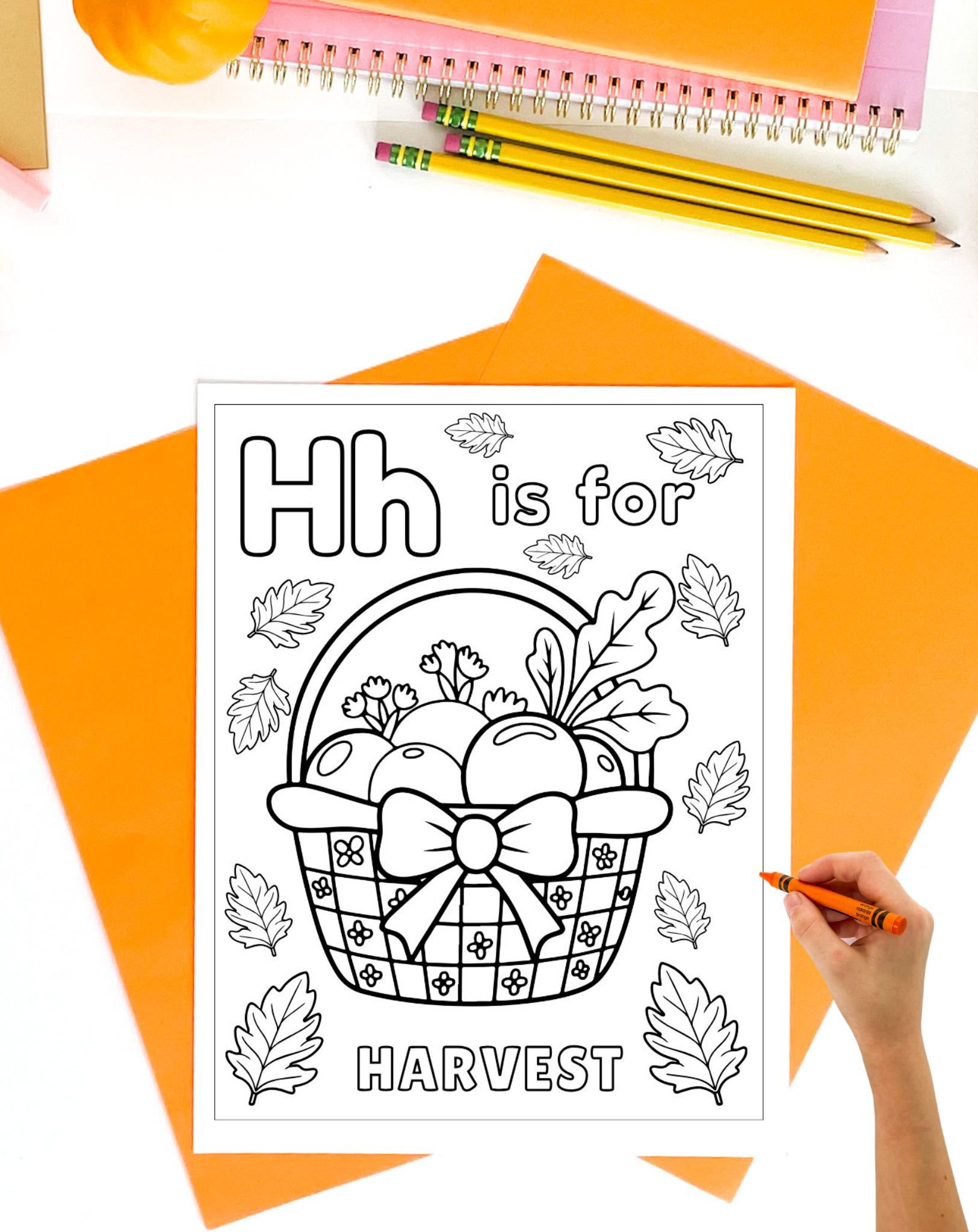 a hand with an orange crayon coloring a page with Hh is for Harvest on it