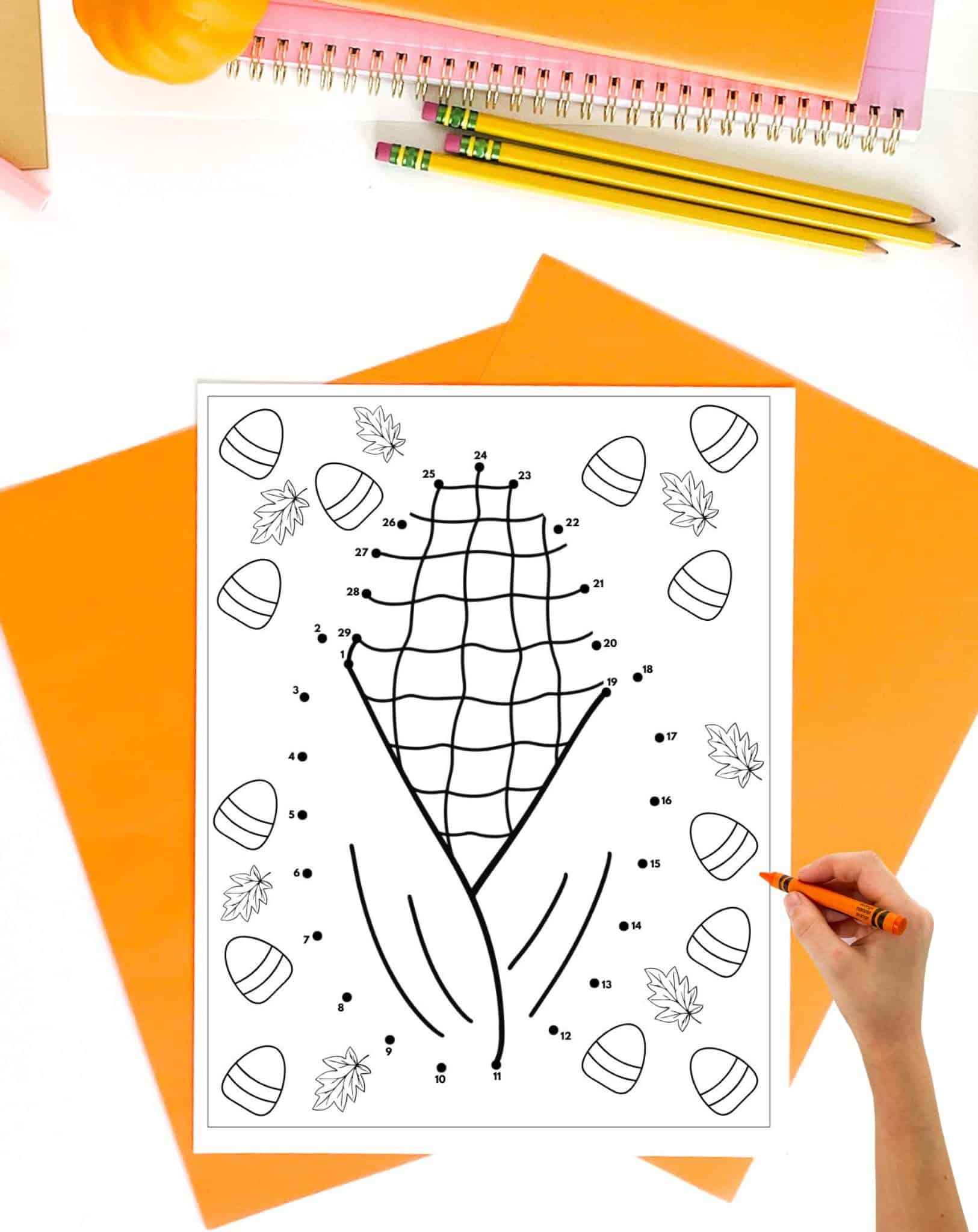A hand is coloring a Thanksgiving-themed connect the dots printable featuring a corn design with numbered dots and surrounding decorative elements like leaves and candy corn, set against an orange backdrop with school supplies.
