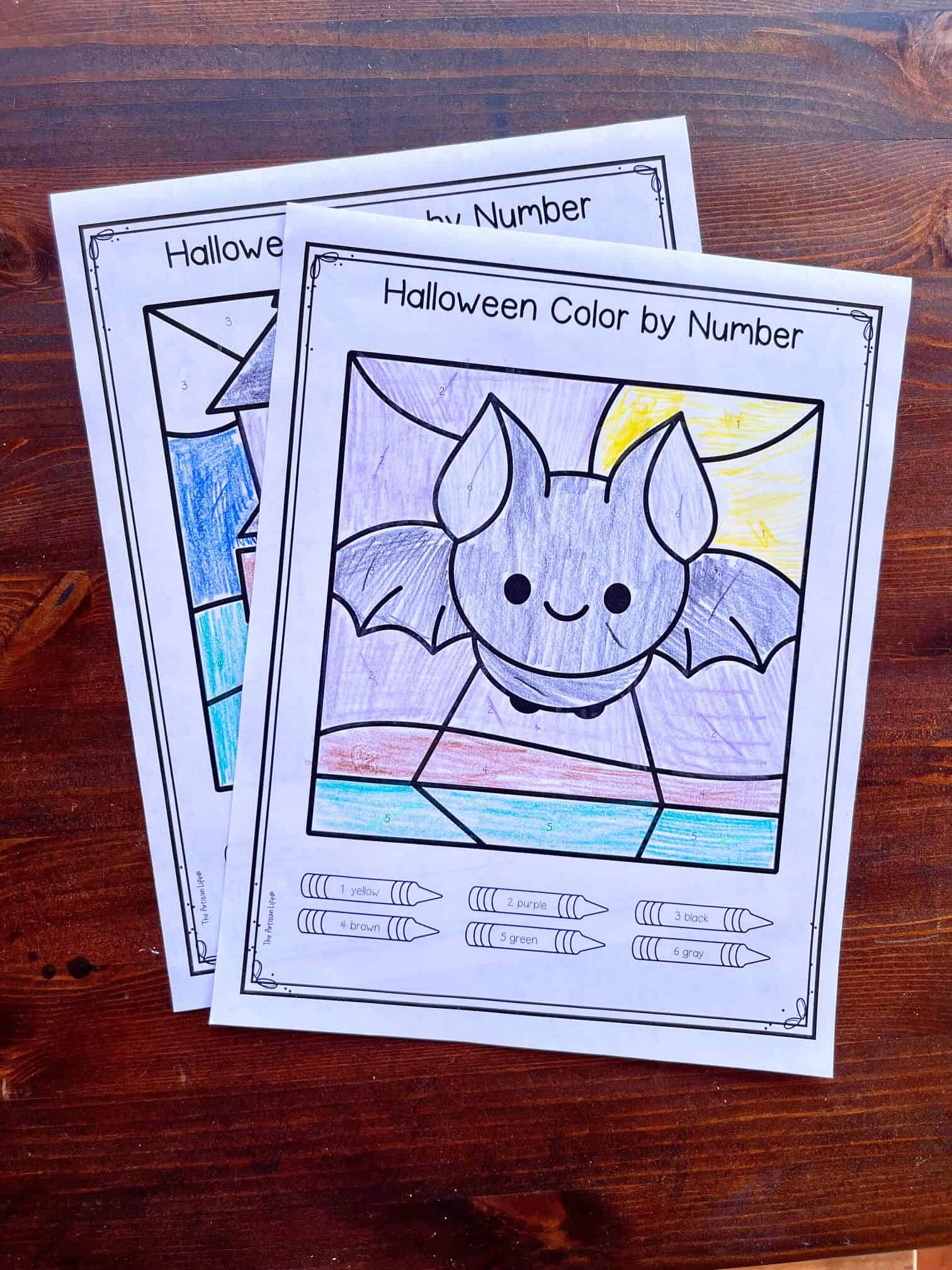 Two Halloween color by number worksheets featuring a bat, partially colored, with crayon color keys for preschoolers on a wooden table.
