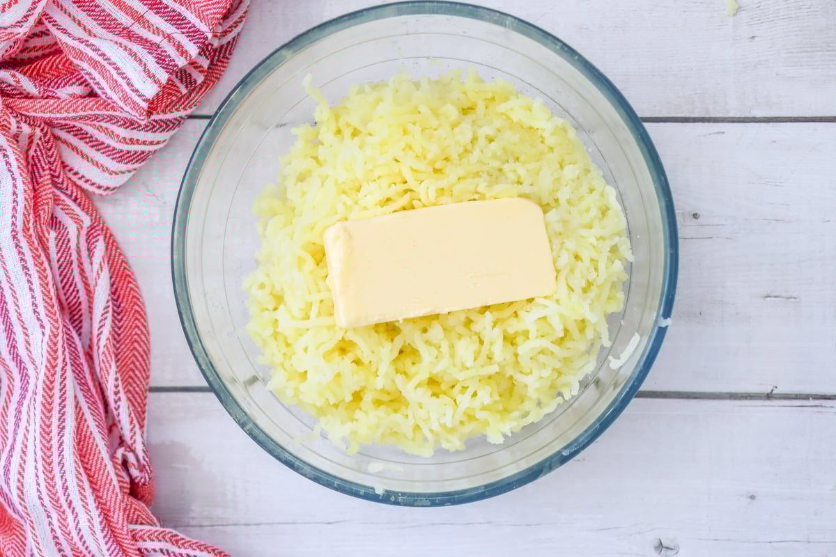 A glass bowl contains freshly mashed potatoes with a stick of butter placed on top, alongside a red and white striped kitchen towel.