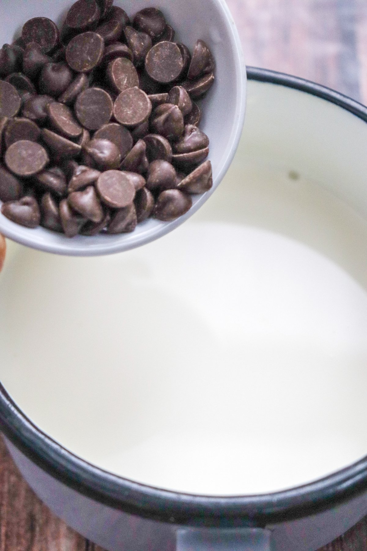 A bowl of chocolate chips is being poured into a pot containing milk, suggesting preparation for a homemade hot chocolate recipe.