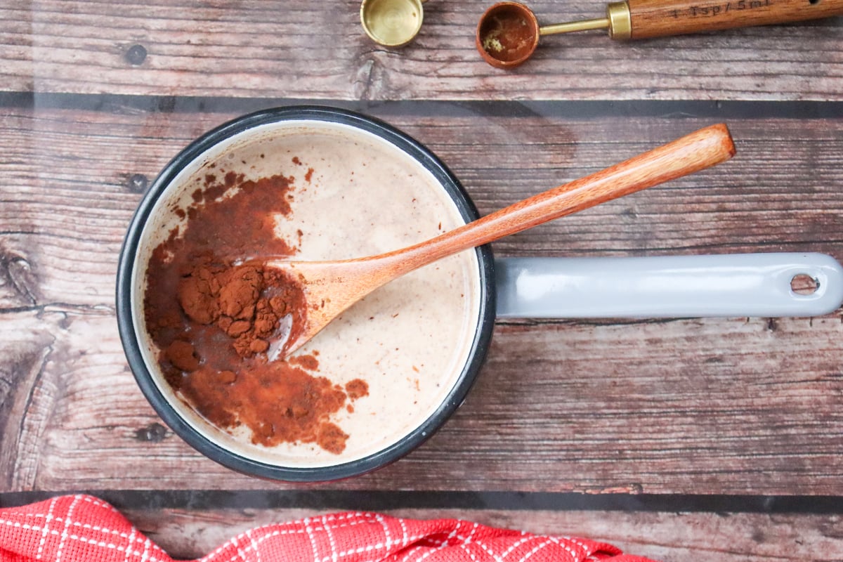A pot of homemade hot chocolate mixture, featuring cocoa powder and a wooden spoon, is set on a wooden surface next to measuring spoons and a red checkered cloth, suggesting an easy DIY recipe for holiday eggnog or chocolate eggnog drinks.