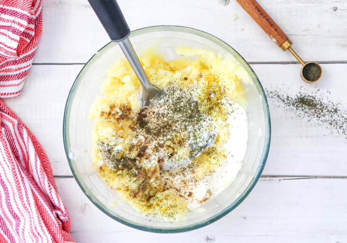 A glass bowl contains creamy mashed potatoes being mixed with seasoning and cream, accompanied by a red and white striped towel and a measuring spoon on a wooden surface, showcasing ingredients for homemade garlic mashed potatoes.