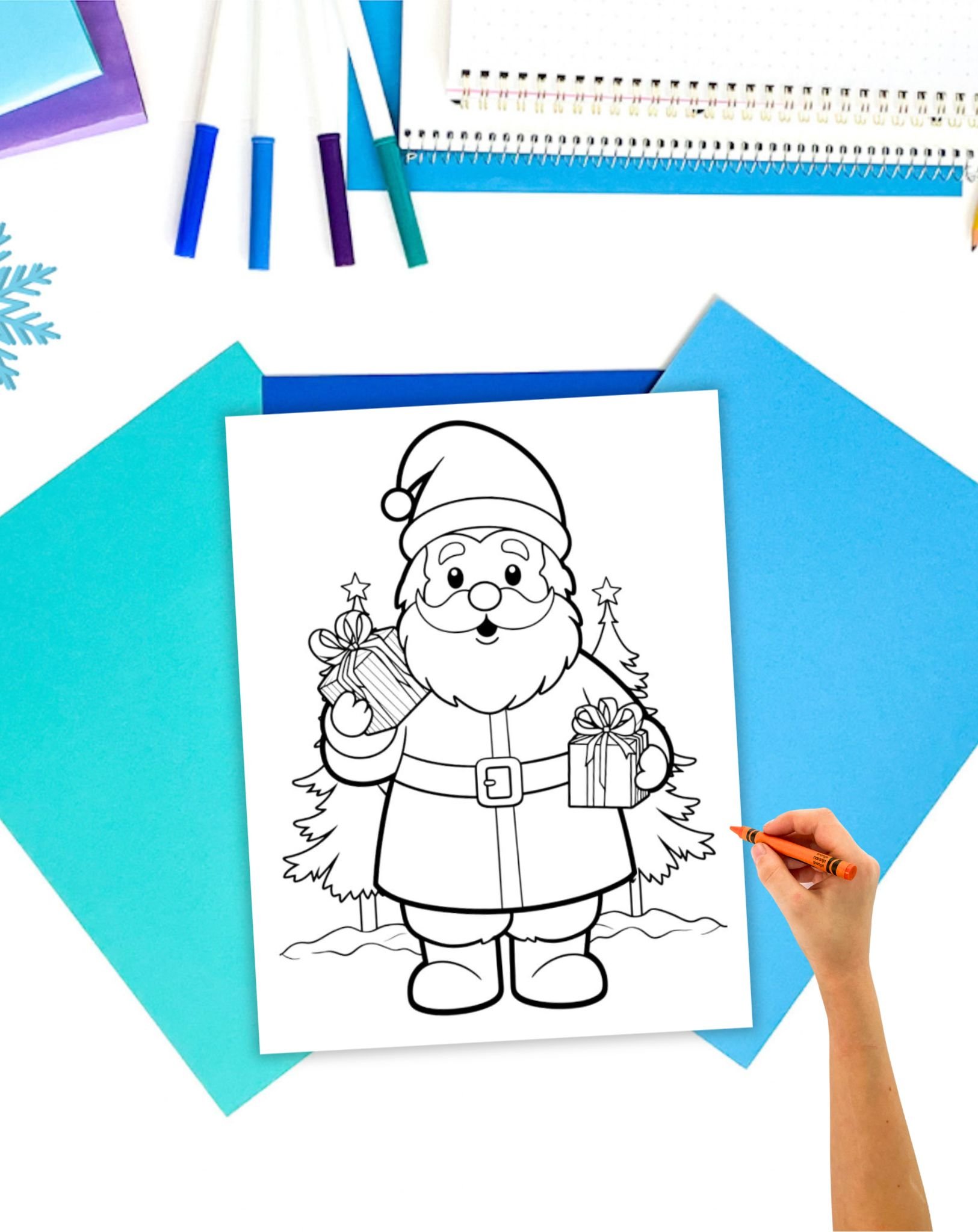 A hand holding a colored pencil is seen next to a Santa Claus coloring page featuring Santa with gifts and Christmas trees, surrounded by vibrant colored paper and stationery.
