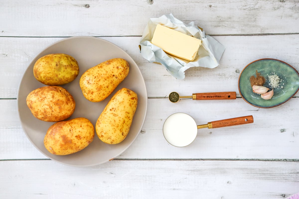 An assortment of ingredients for making creamy mashed potatoes is displayed, including five raw potatoes on a plate, a stick of butter wrapped in paper, measuring spoons, milk, and garlic on a small dish.