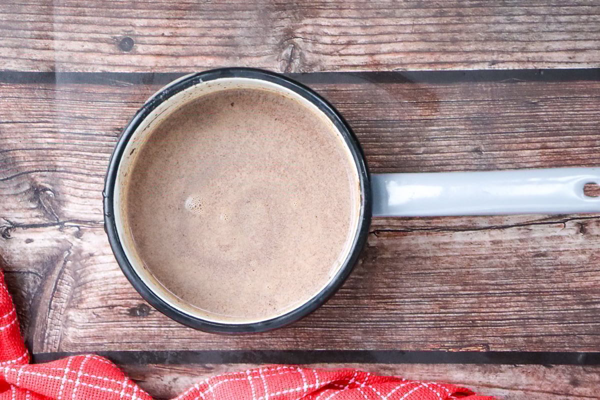 A saucepan containing homemade hot chocolate sits on a wooden surface next to a red checkered cloth, suggesting a cozy atmosphere for enjoying easy homemade hot chocolate or holiday eggnog drinks.