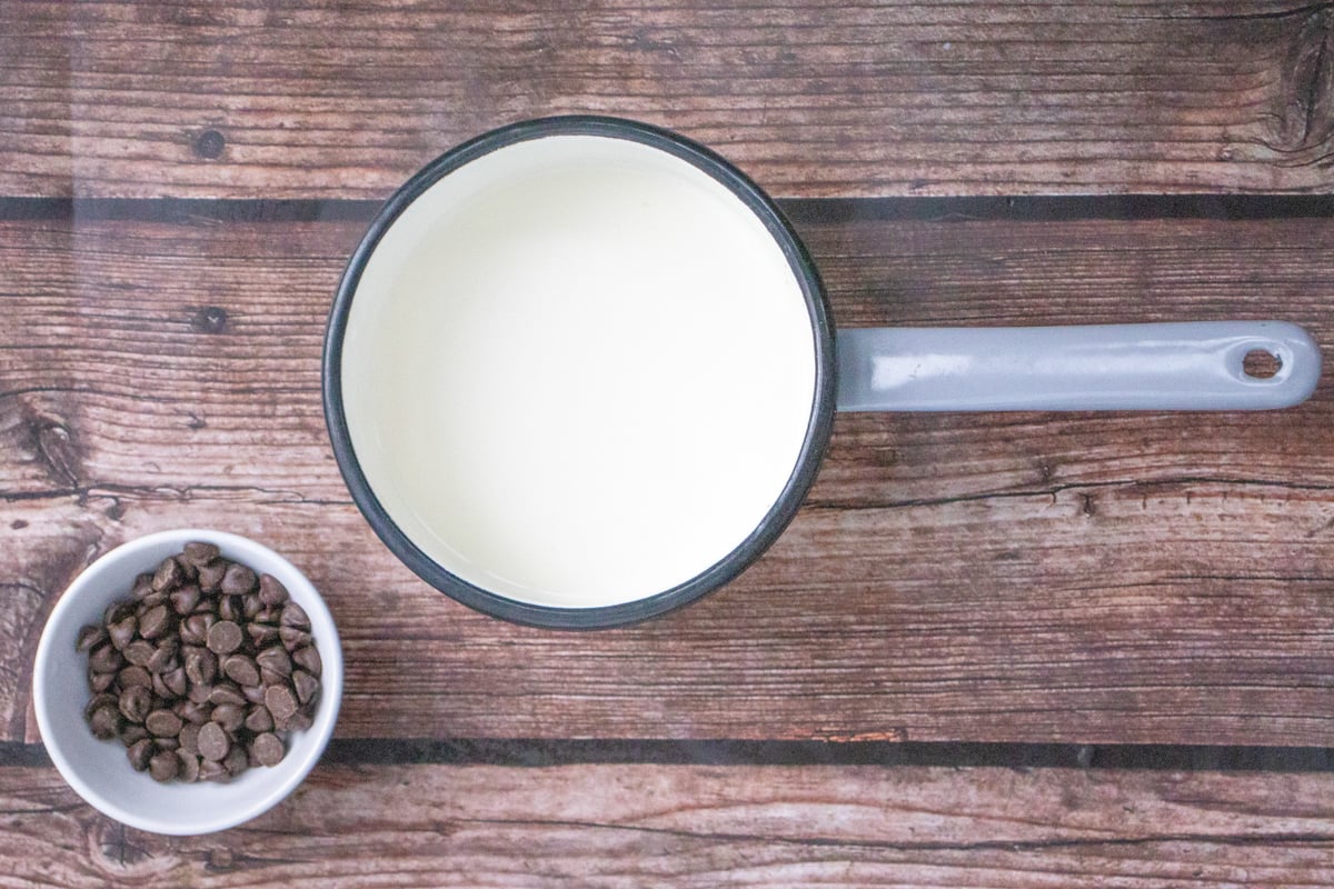 A saucepan with warm milk sits next to a small bowl of chocolate chips, suggesting preparation for a homemade hot chocolate recipe.