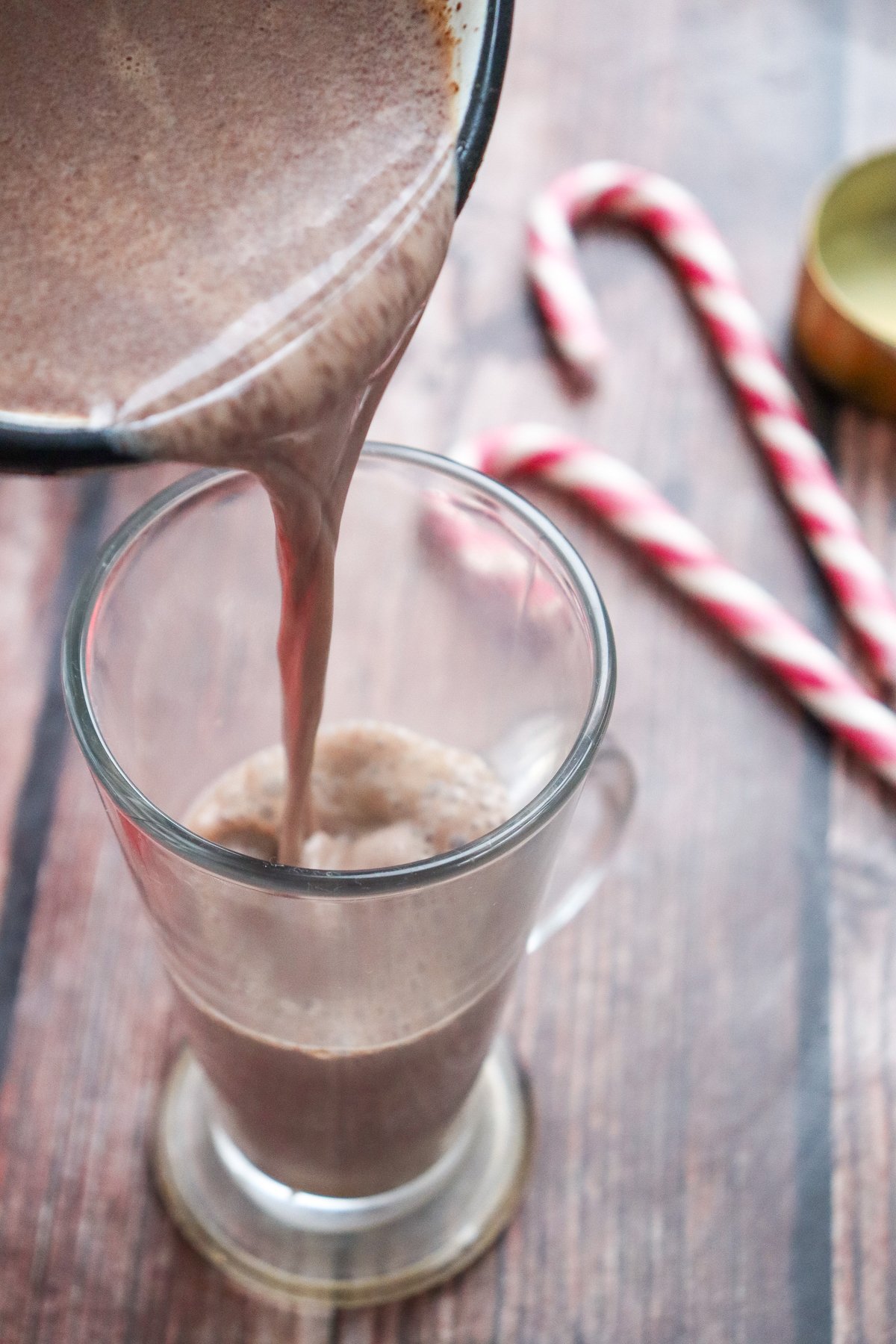 A homemade hot chocolate is being poured from a saucepan into a clear glass, with candy canes visible in the background.