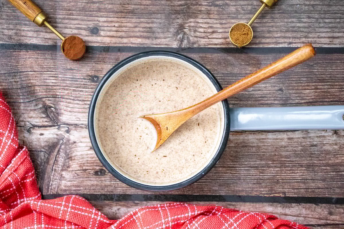 A pot of homemade hot chocolate sits on a wooden table, with a wooden spoon inside and two measuring spoons nearby, one containing cocoa powder.