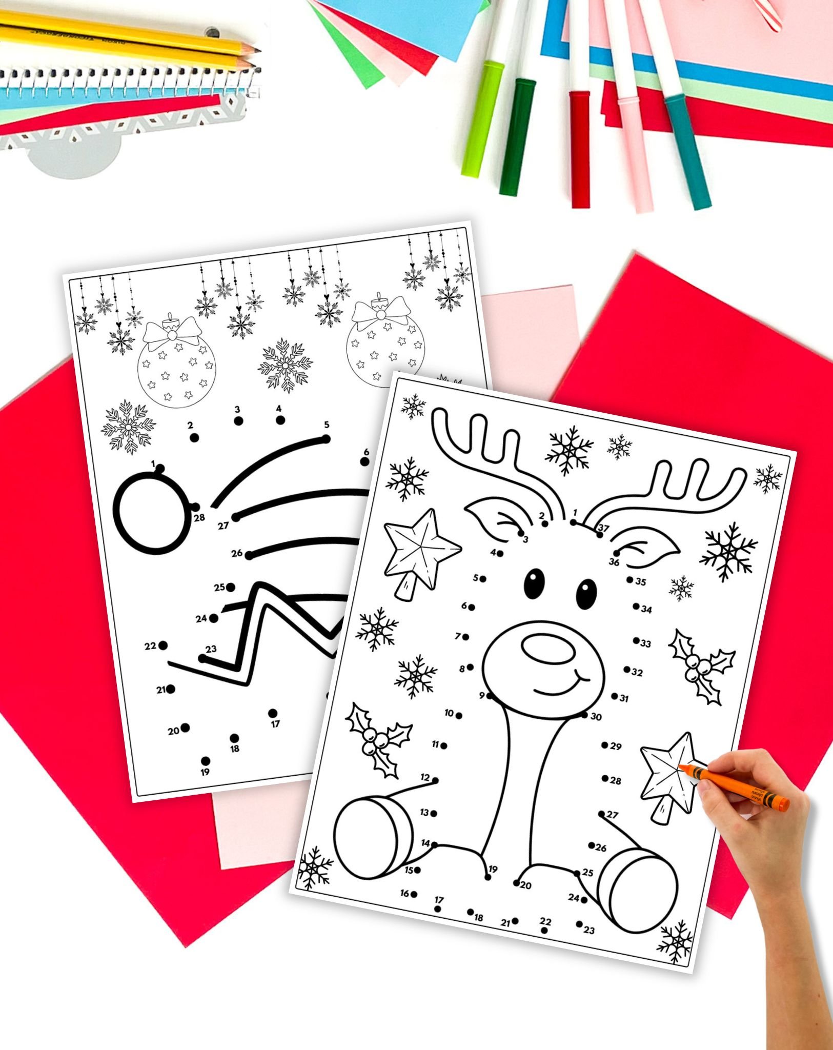 Two Christmas connect the dots worksheets on a table with colorful papers and markers. A child's hand prepares to color the images. 
