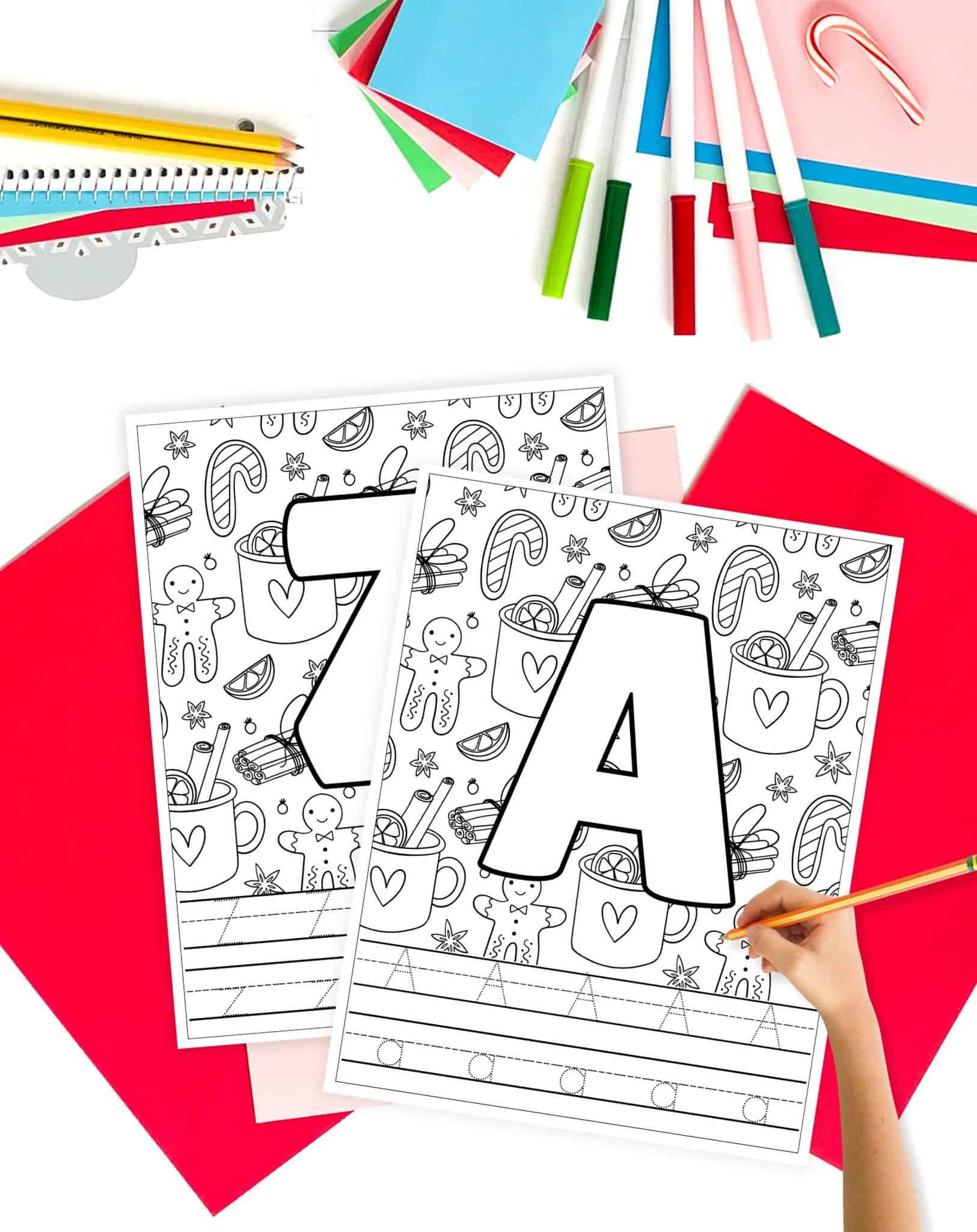 letters a and z tracing pages with christmas elements to color. The pages are on a table with christmas colored papers and a hand holding a pencil is preparing to trace the worksheets