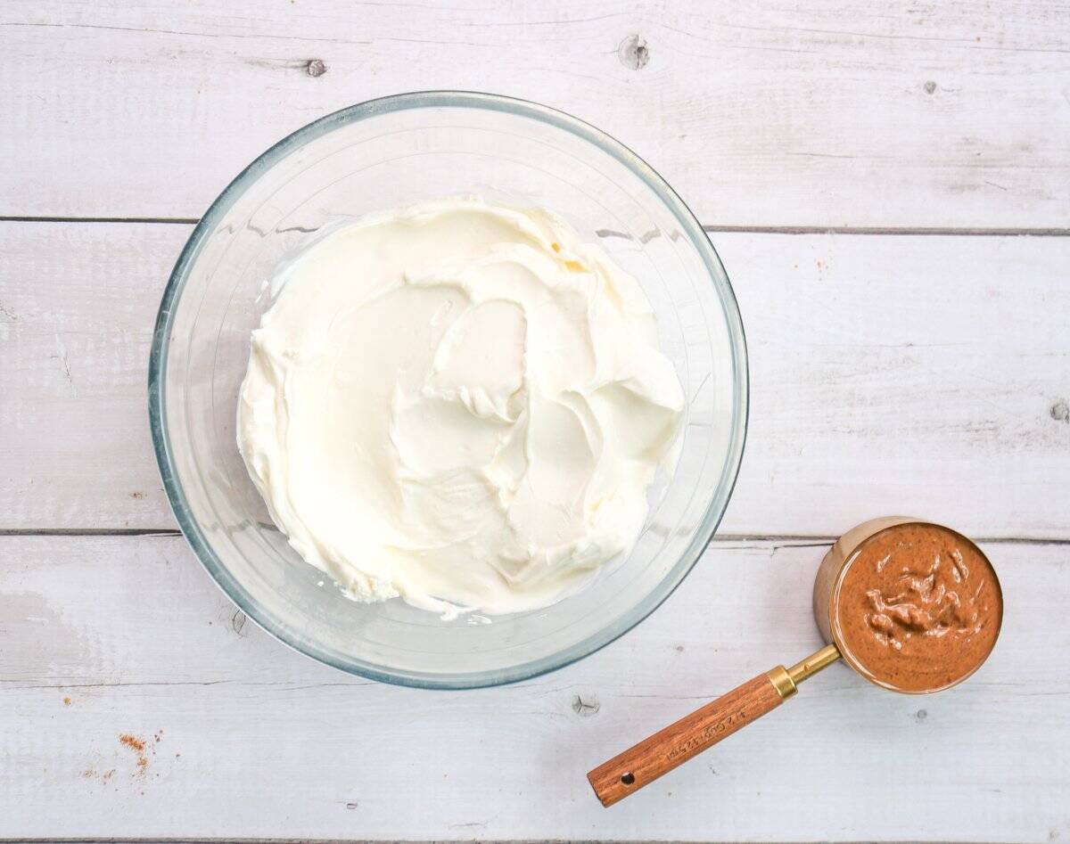 A glass bowl filled with cream cheese sits on a wooden surface alongside a measuring cup containing peanut butter