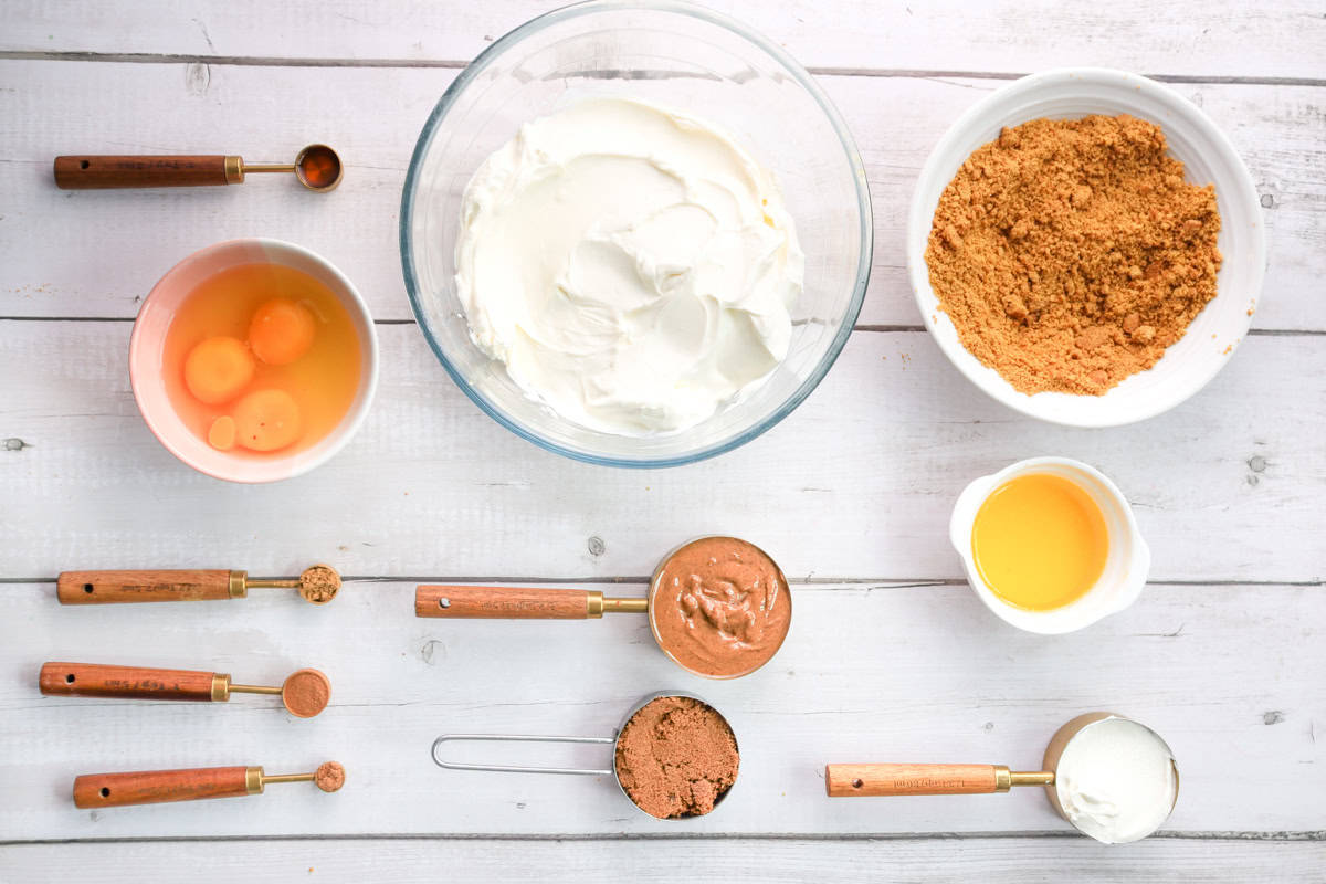 Ingredients for a gingersnap cheesecake, including cream cheese, eggs, gingerbread mixture, and crushed gingersnap cookies, arranged on a wooden surface, reflecting preparations for holiday baking recipes.