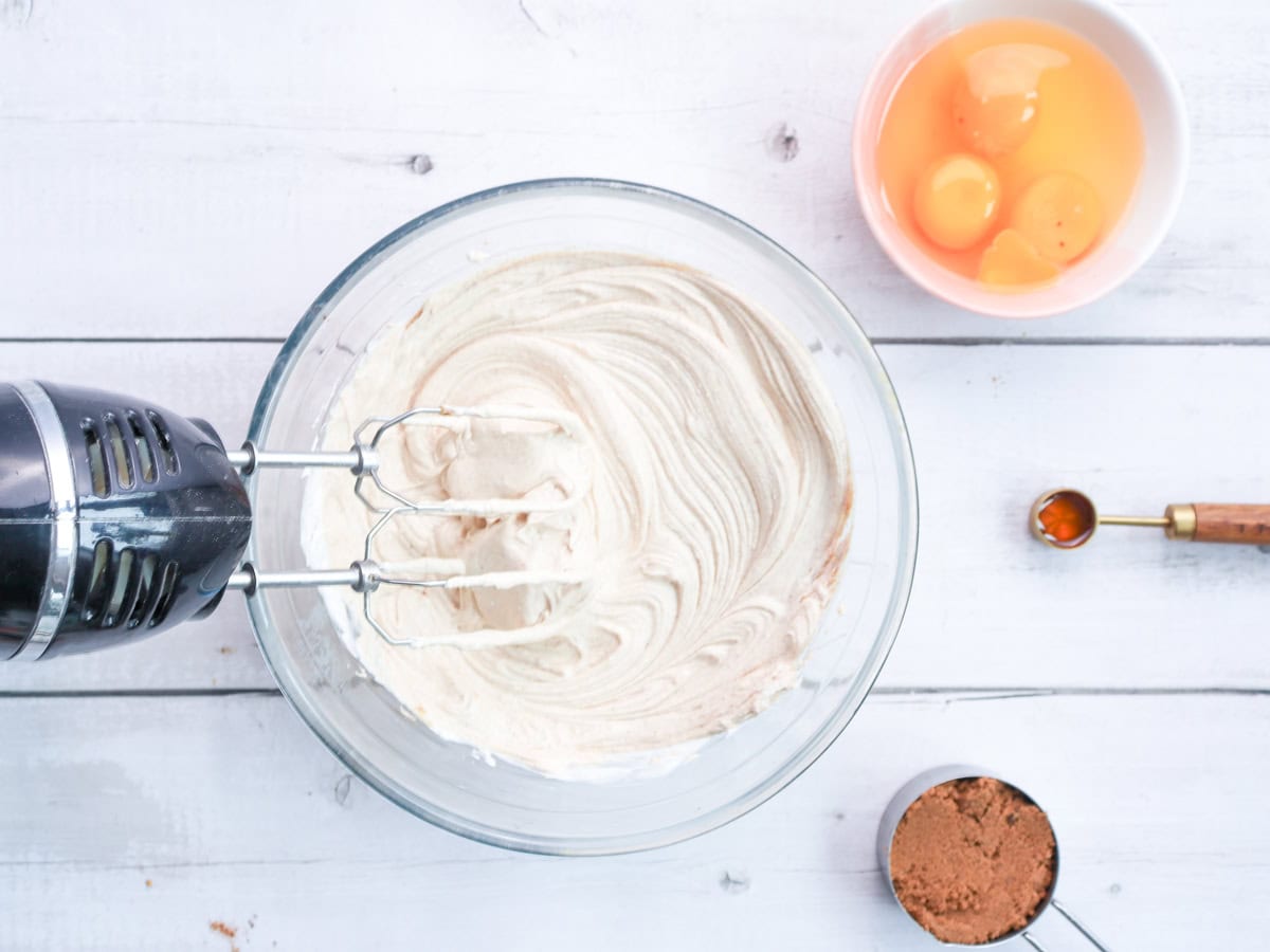 A mixing bowl with a blended cheesecake mixture sits next to a bowl of eggs, a measuring spoon, and a cup of brown sugar, highlighting the preparation of a gingerbread cheesecake or gingersnap cheesecake for holiday baking.