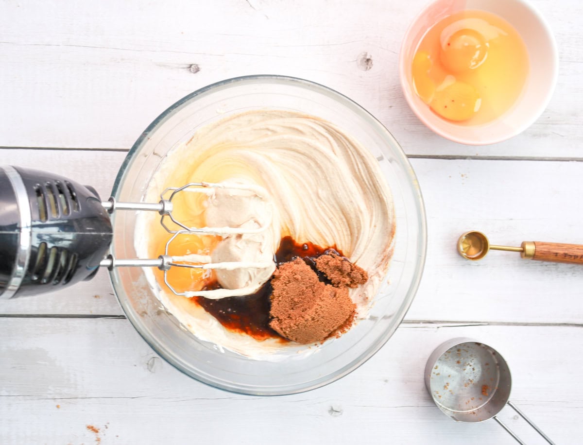 A mixing bowl contains a blend of cream cheese, brown sugar, and eggs, with a hand mixer and measuring tools nearby, suggesting preparation for a gingersnap cheesecake or gingerbread cheesecake for holiday baking recipes.