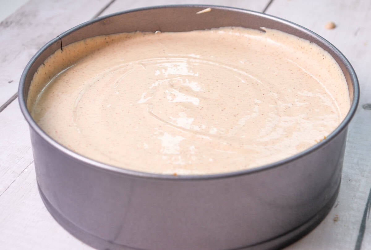 A round cake pan filled with unbaked gingersnap cheesecake batter sits on a wooden surface, ready for holiday baking.