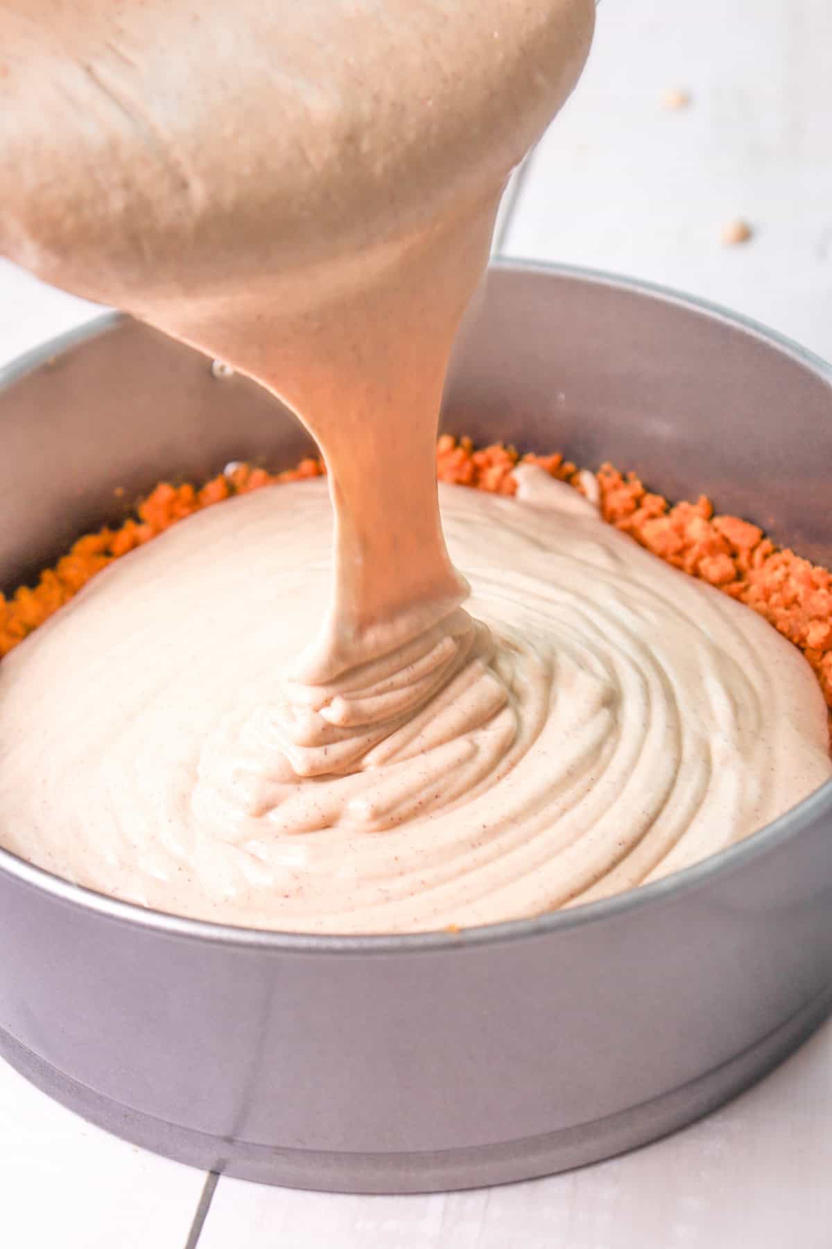 A thick, creamy mixture is being poured into a springform pan with a gingersnap crust, preparing to make a gingerbread cheesecake for holiday baking.