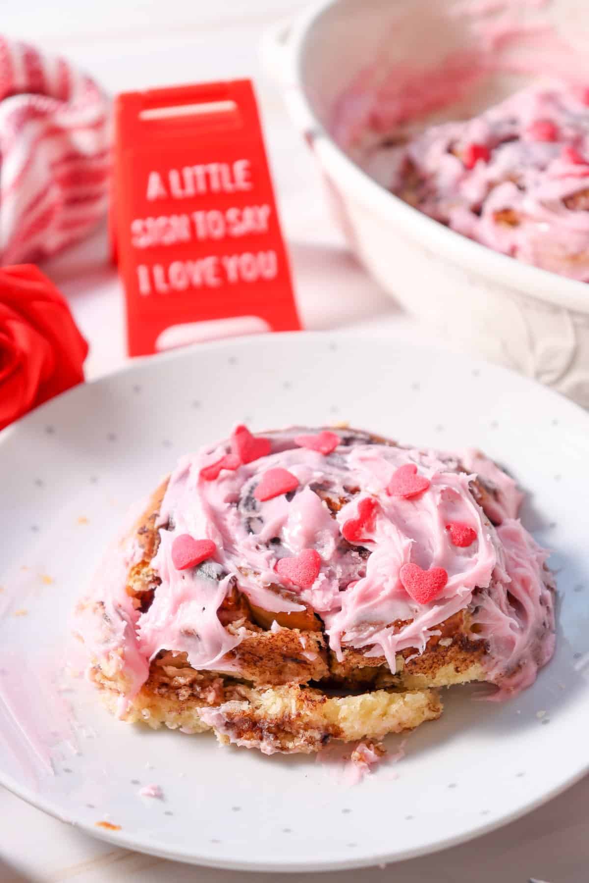 A close-up of a homemade cinnamon roll topped with pink frosting and heart-shaped sprinkles, with a sign in the background that reads "A little sign to say I love you," representing easy small batch cinnamon rolls perfect for Valentine's Day.