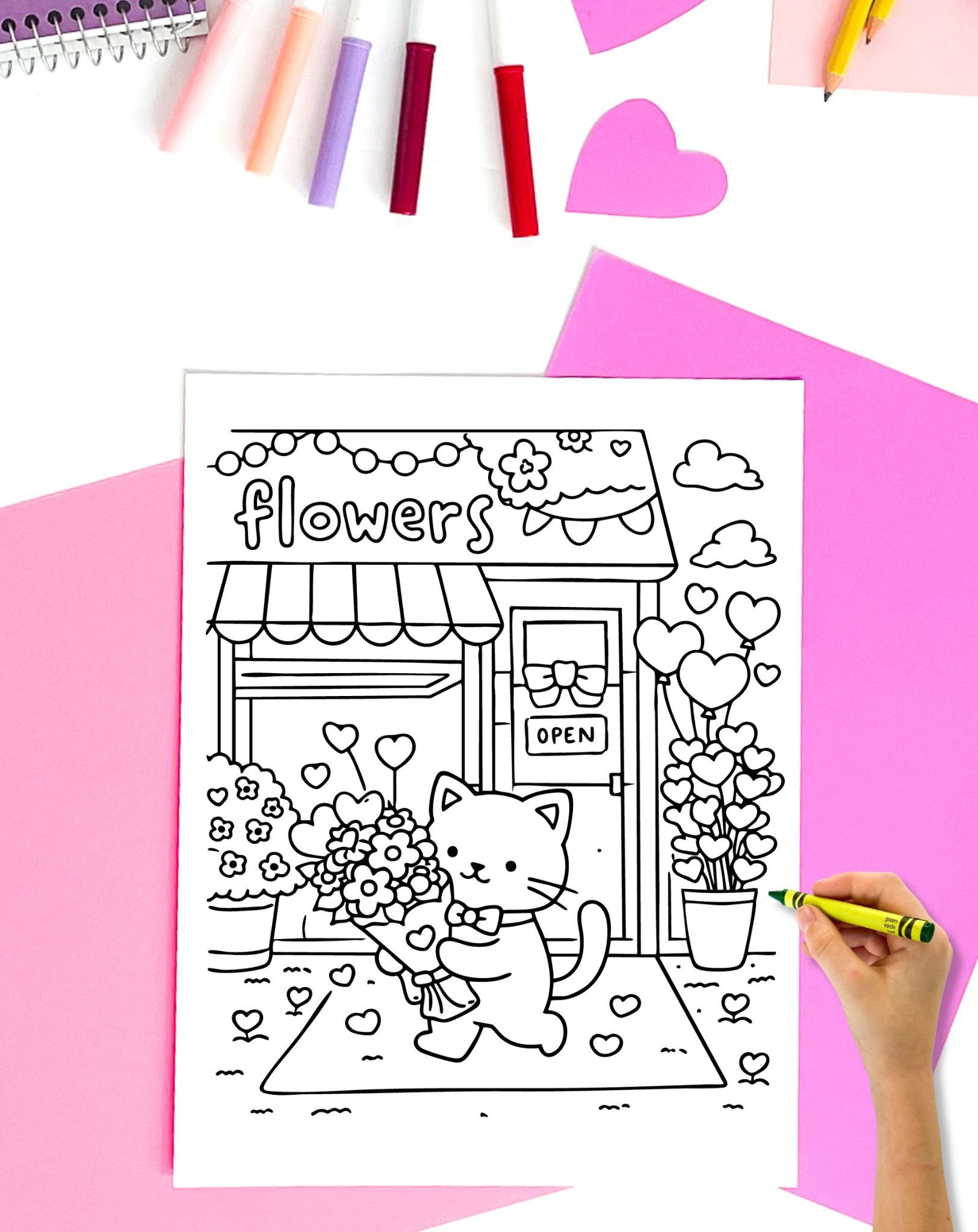 a child's hadn't with a green crayon preparing to color a Valentine's Day coloring page showing a cat with flowers