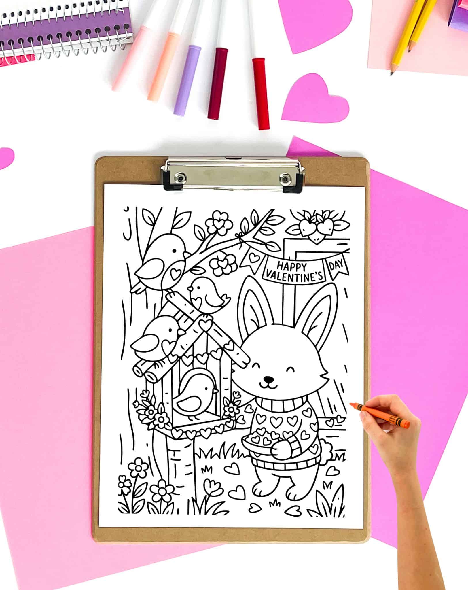 a hand with a crayon preparing to color a cute Valentine's Day coloring page that's on a clipboard