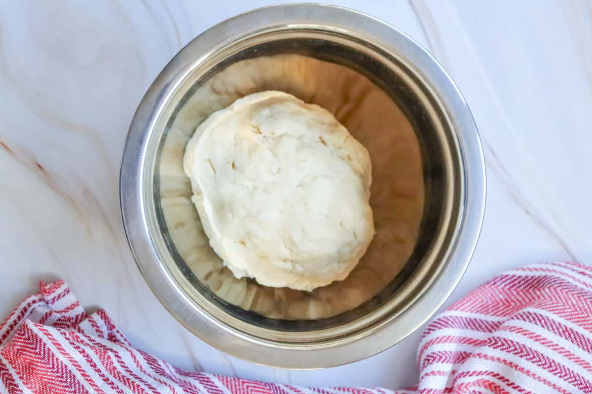 A metal bowl contains a lump of dough prepared for making easy small batch cinnamon rolls, with a red and white striped towel visible nearby, suggesting a homemade treats context.