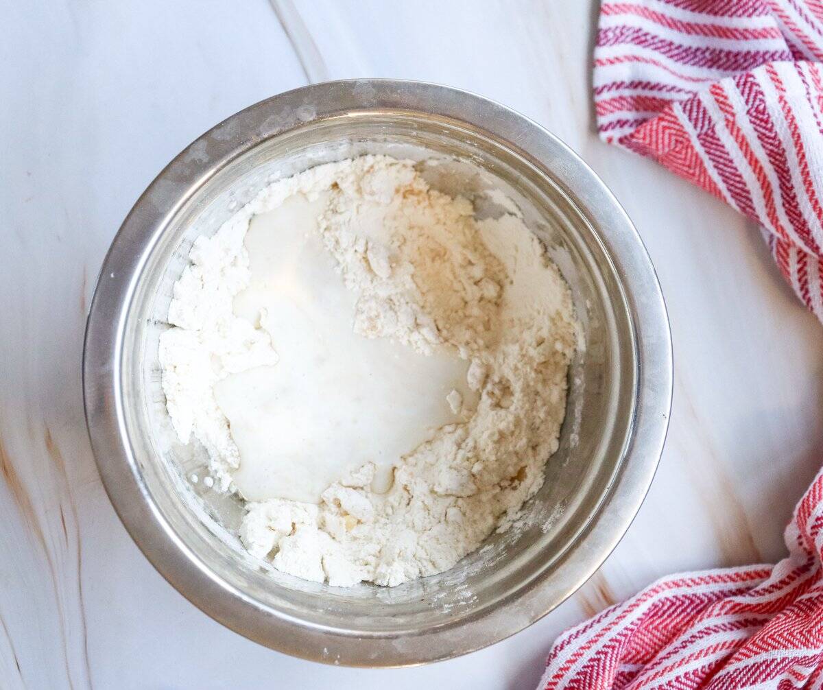 A stainless steel bowl contains a mixture of flour and liquid ingredients, preparing for an easy small batch cinnamon roll recipe, with a red and white striped dish towel visible nearby.