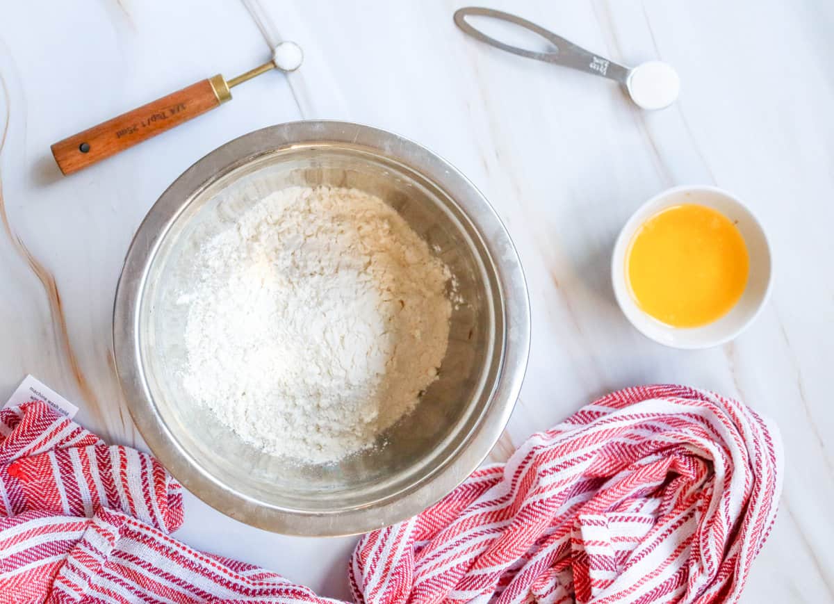 A metal bowl containing flour is centered on a marble surface, accompanied by an egg in a small dish, a measuring spoon, and a red and white striped kitchen towel, suggesting preparation for an easy small batch cinnamon rolls recipe.