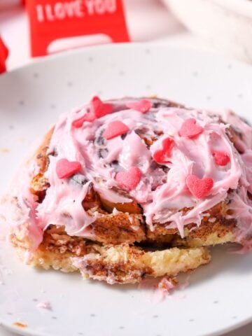 A close-up of a cinnamon roll topped with pink frosting and heart-shaped sprinkles, placed on a dotted plate, next to a red "I Love You" card, showcasing a homemade treat ideal for Valentine's Day and featuring a small batch cinnamon roll recipe.