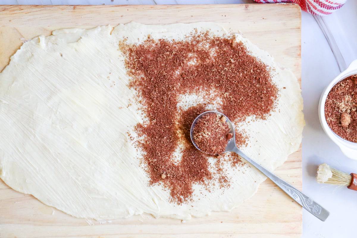 A wooden cutting board displays a rolled sheet of dough with cinnamon sugar sprinkled on top, accompanied by a small bowl and brush, illustrating the process of making easy small batch cinnamon rolls.
